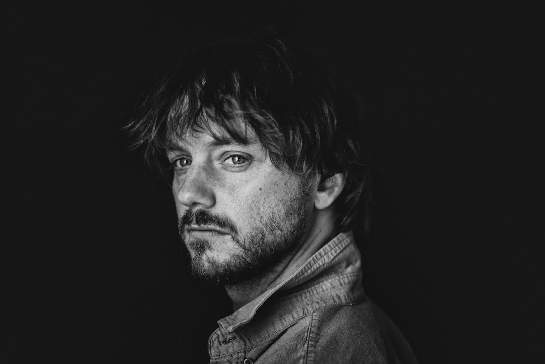 A man with dark, messy hair and a beard, wearing a denim jacket, stands in a dimly lit room.