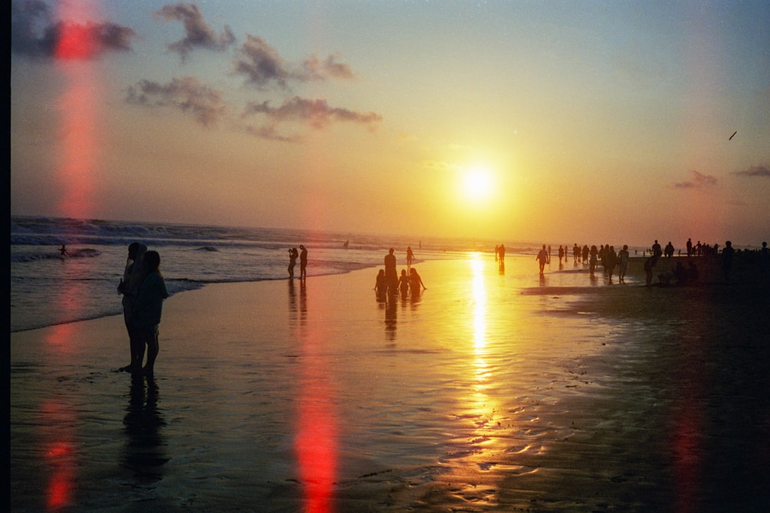A vibrant sunset casts a warm glow on the beach, where people stroll and play in the shallow water.