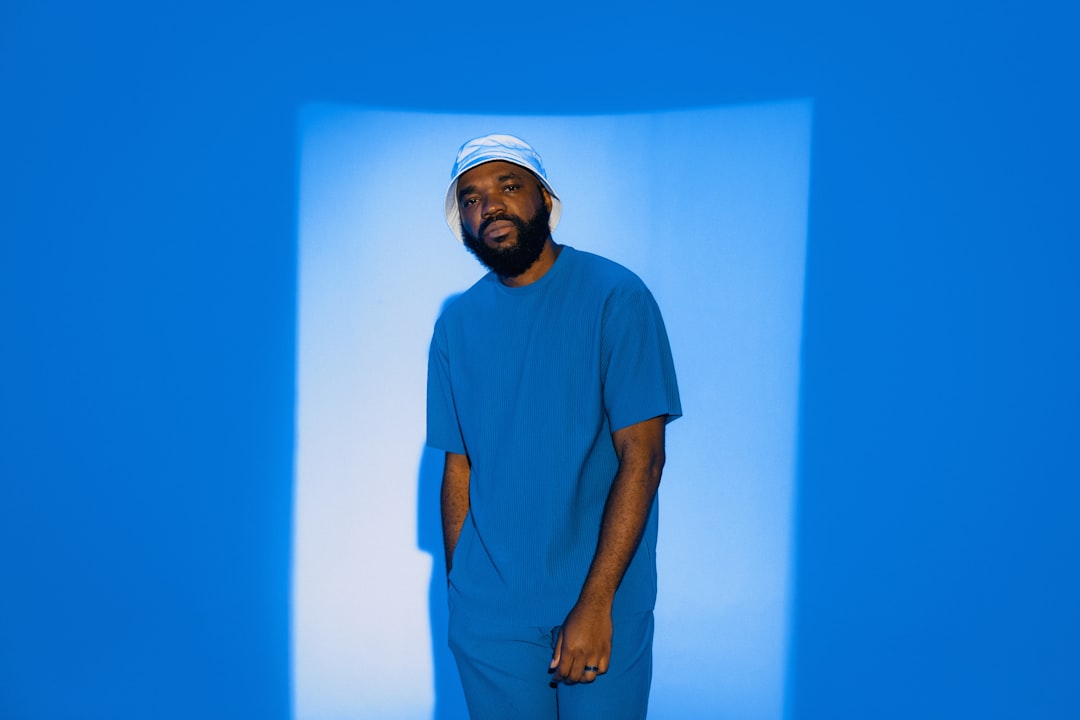 A man in a blue shirt and matching pants stands against a blue wall, illuminated by a beam of light.