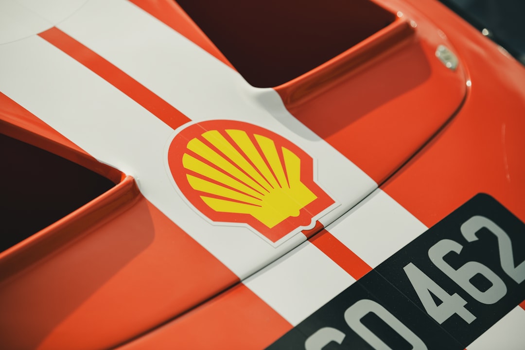 A vibrant orange race car with a prominent yellow shell logo, adorned with white stripes, is captured in a close-up shot.