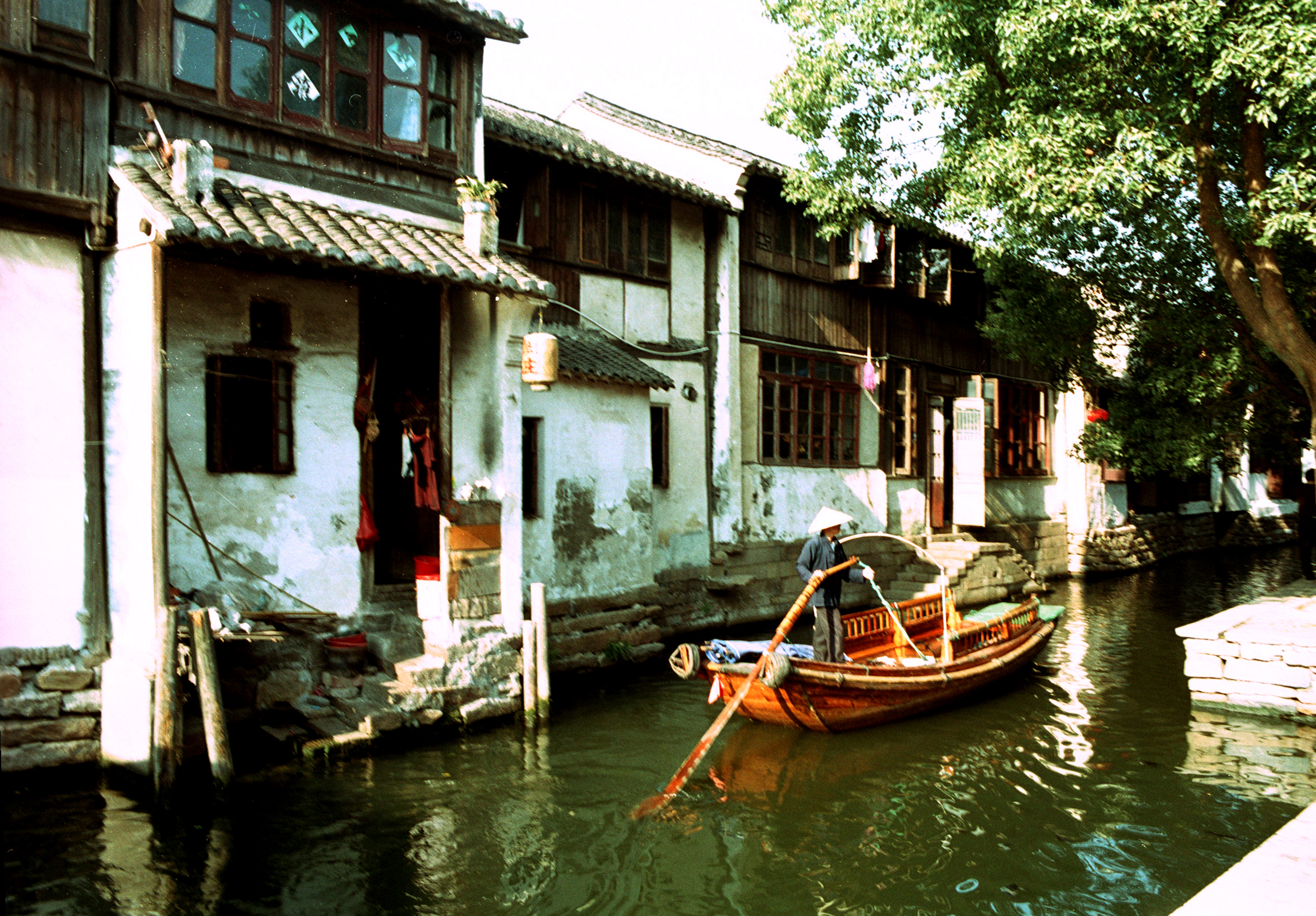 A small boat navigates a serene canal lined with traditional buildings, surrounded by lush greenery.