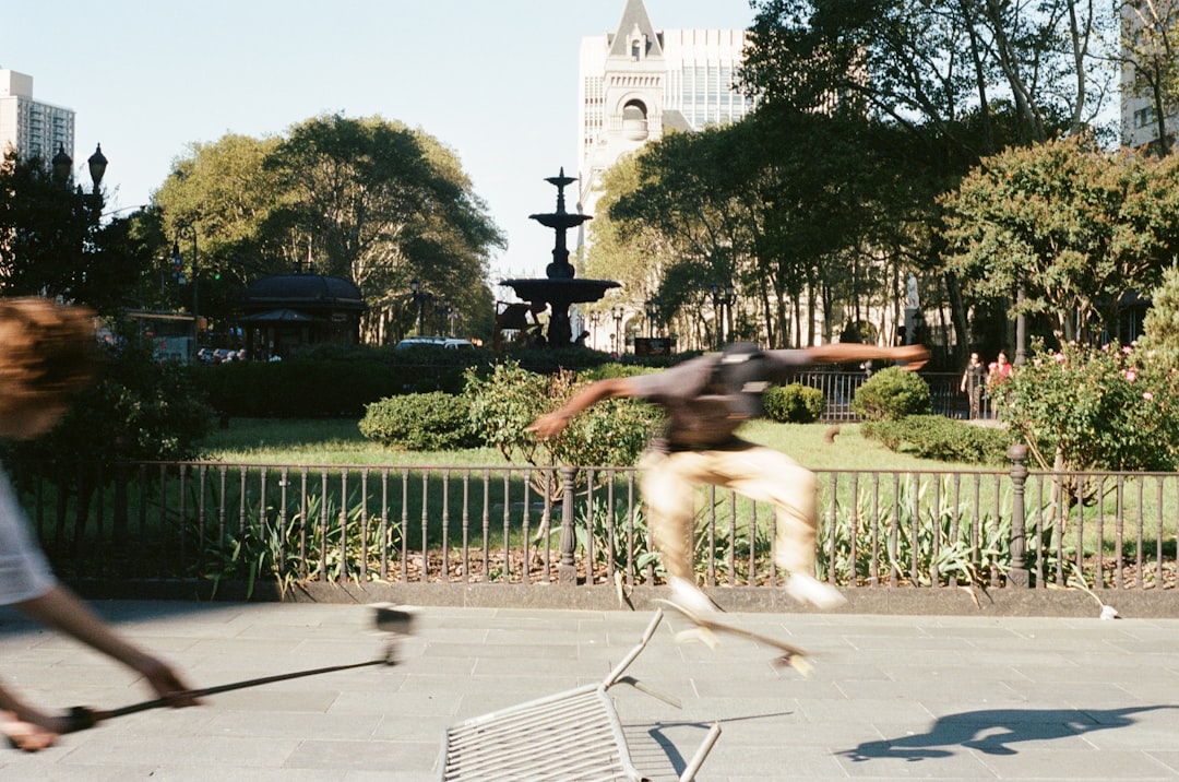 A man in a grey shirt and khaki pants leaps into the air, his arms outstretched, in front of a fountain.