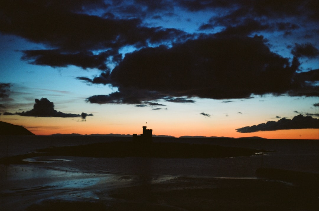 A castle stands silhouetted against a vibrant sky, with clouds and a sunset unfolding behind it.