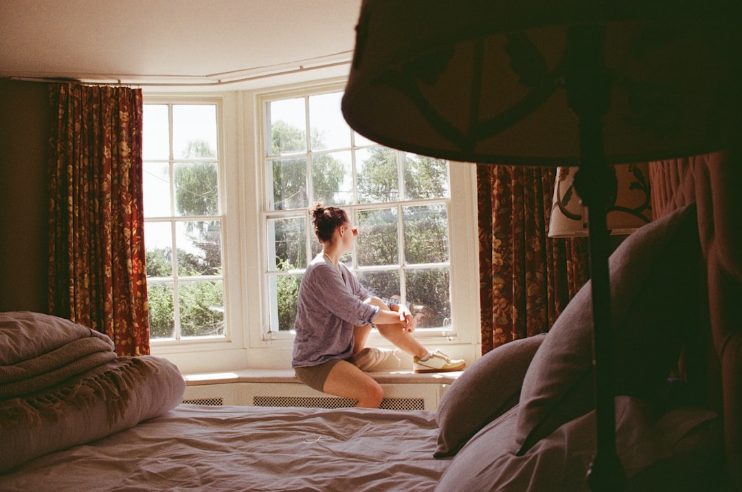 A woman sits on a windowsill, gazing out at trees and bushes through a large window.