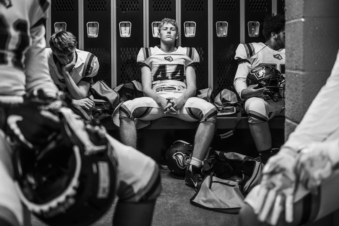A young football player sits on a bench in a locker room, wearing a white uniform with the number 44 on his jersey.