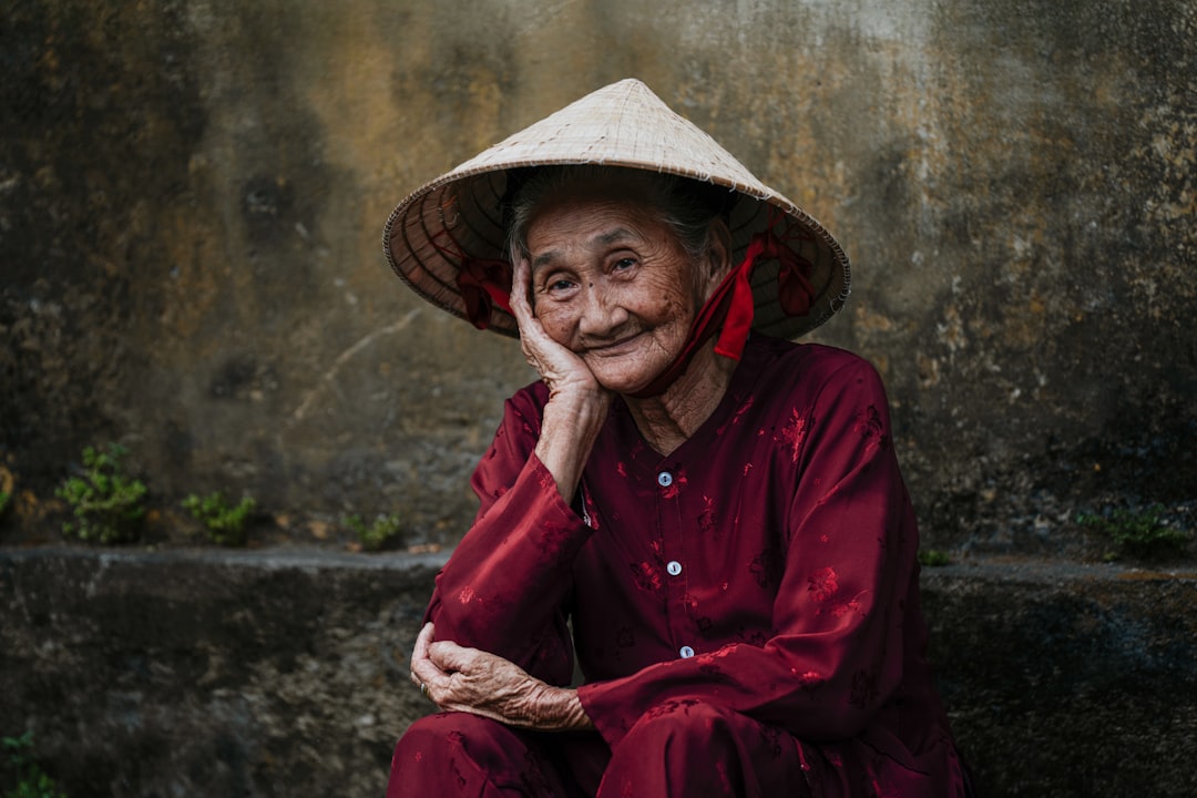 An elderly woman in a red outfit and conical hat sits in front of a weathered stone wall.