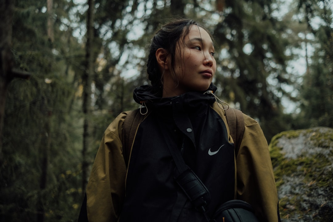 A woman stands in a forest, her dark hair pulled back, wearing a black Nike jacket with a white logo, and carrying a black bag.