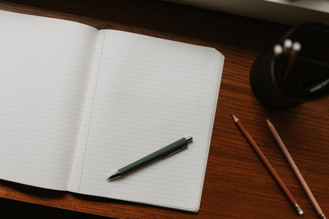 A green pen lies on an open notebook, surrounded by pencils on a wooden desk.