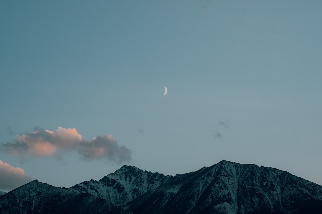 A serene mountain range is set against a soft blue sky, with a crescent moon shining brightly.