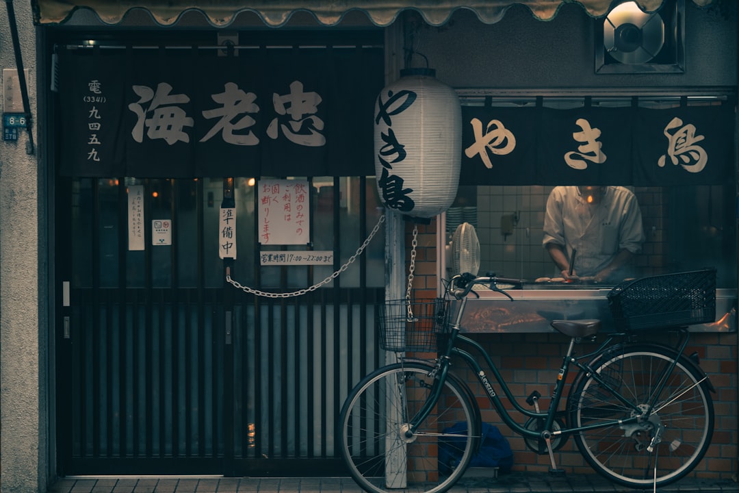 A dark storefront with Japanese writing and a bicycle parked outside, featuring a chef in a white shirt working at a counter.